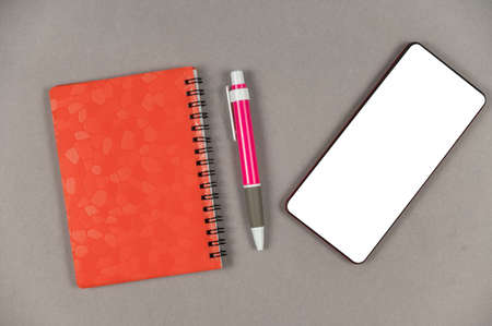 Notepad, pen and smartphone on gray background. Red closed spring-loaded notebook. plastic pen. Smartphone with white blank screen. Copy space for text and design elementsの写真素材