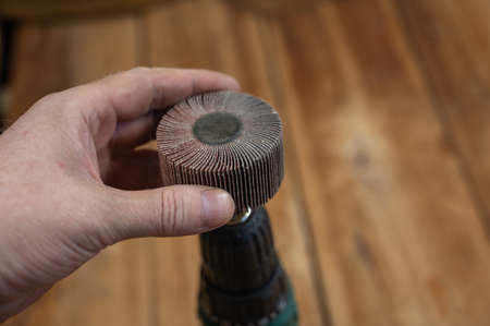 Man places flap wheel in cam of an electric drill chuck. Tool for woodworking, sanding, removing rust from various surfaces. Yellow wooden boards defocused in background. close-up. selective focus.の写真素材