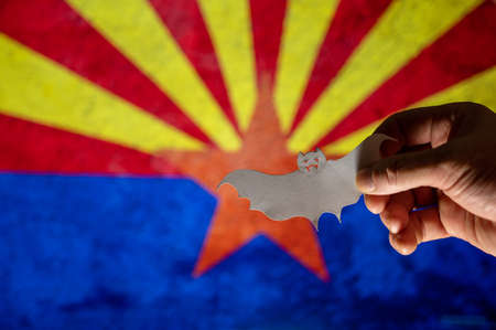 Happy Halloween, Arizona. Male hand holding bat figurine. Arizona state flag in background. Cut out white paper Halloween paraphernalia. All Hallows Evening. selective focus.の写真素材
