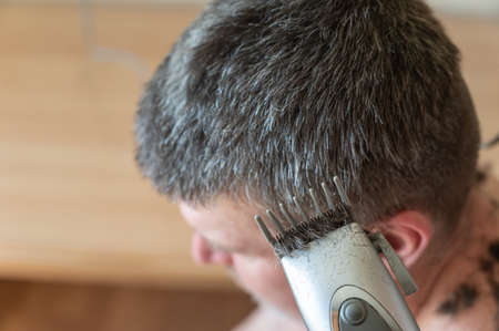 Wife cuts her husband's hair at home. A woman uses an electric hair clipper to cut short, graying hair. Middle-aged person with a naked torso sits inside a living room. front focus.の写真素材