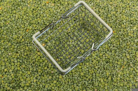 An empty shopping basket stands on top of dry chopped peas. A small metal shopping basket on top of a surface strewn with green uncooked beans. close-up. selective focus.の写真素材