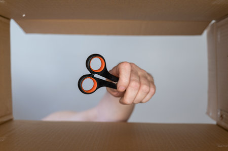 A man's hand places stationery scissors in a cardboard moving box. Hand tool for cutting paper and other thin objects. bottom view. inside view. close-up. selective focus.の写真素材