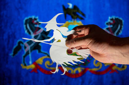 Witch flying on broomstick in front of Commonwealth of Pennsylvania flag. Man hand holding figurine cut out of paper. Celebrating All Saints Night in United States concept. selective focus.の写真素材