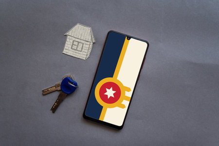 Bunch of door keys, a paper house, and a smartphone on a gray background. Tulsa city flag on the screen of the device. Buying real estate in Tulsa, Oklahoma, USAの写真素材