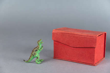 Red box and miniature of one dinosaur against a gray background. Small green figurine of an animal of prey standing on its hind legs. Rectangular closed box. selective focus.の写真素材