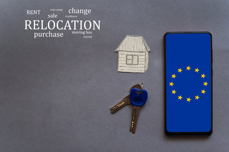 Change of residence concept. Smartphone, paper figurine of house, door keys. EU flag on smartphone screen. Buying or selling real estate in Europe. Themed words at the top left of the picture.の写真素材