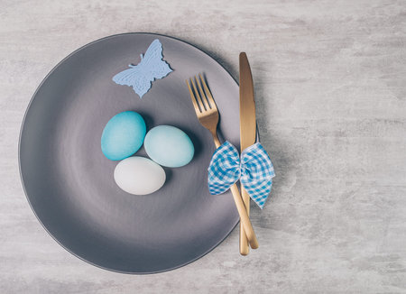 Easter table setting with grey plate and blue eggs on grey wooden table. 