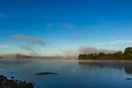 Fishermen on boats early in the morning at dawn in the fog at golden hour catch fish on the Oka River, Russia. Barge, tugboat, green grass and trees, a river bend against a clear light sky in summer.の写真素材