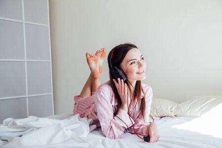 brunette in pink Pajamas listening to music with headphones on the bedの写真素材