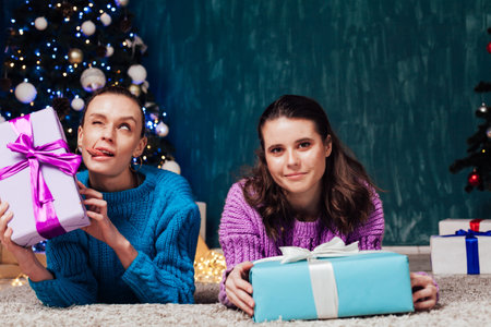 Two beautiful female girlfriends open Christmas gifts at the Christmas tree for the New Year holidayの写真素材