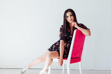 Beautiful fashionable brunette woman in a dress with flowers sits on a chair in the room