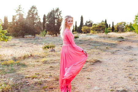 Beautiful fashion blonde woman in pink dress walks in the park at sunsetの写真素材