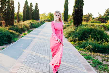 Beautiful fashion blonde woman in pink dress walks in the park at sunsetの写真素材