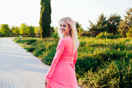 Beautiful blonde woman in pink fashionable dress on a walk in the park at sunsetの写真素材