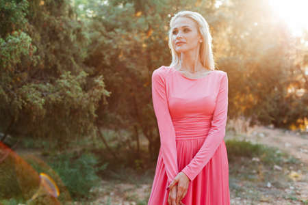 Beautiful blonde woman in pink trendy portrait dress in the park at sunsetの写真素材