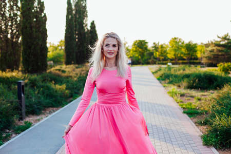 Beautiful blonde woman in pink dress walks in the park at sunsetの写真素材