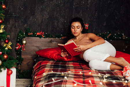 Beautiful woman reads a book on the bed near the Christmas tree New Yearの写真素材