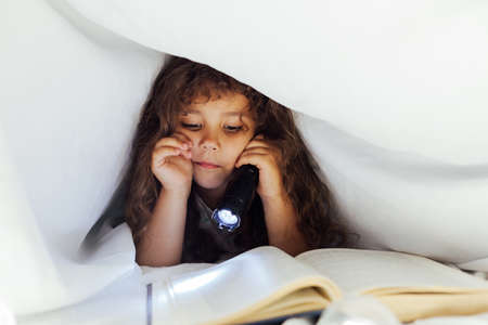 little girl reading a book under a blanket with a flashlightの写真素材