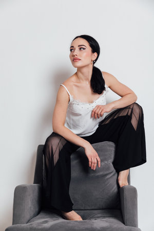 a fashionable woman in a white T-shirt and black pants sits on a chair with a white backgroundの写真素材