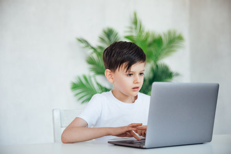 a child sits with a laptop at a table in a room with a green plantの写真素材