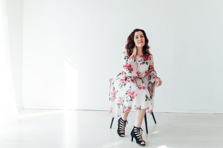 a brunette woman in a floral dress sits in a chair on a light backgroundの写真素材