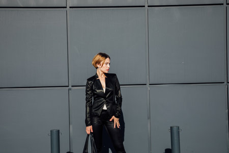 a fashionable woman in leather black clothes stands at a textured black wallの写真素材
