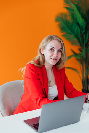 Attractive cheerful business woman in red shirt working on laptop at modern officeの写真素材