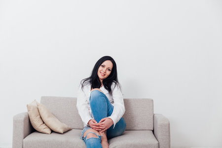 Beautiful brunette woman sitting on grey sofa in white roomの写真素材