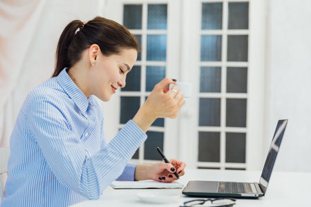 Image of young pleased happy cheerful cute beautiful business woman sit indoors in office using laptopの写真素材