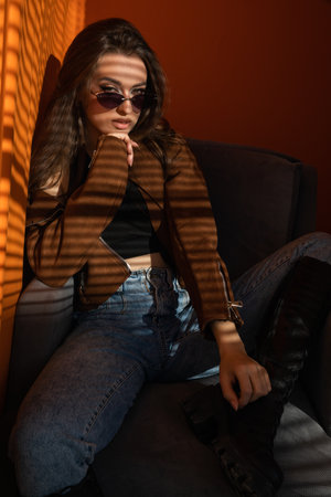 Woman wearing sunglasses posing sitting on orange background at sunsetの写真素材