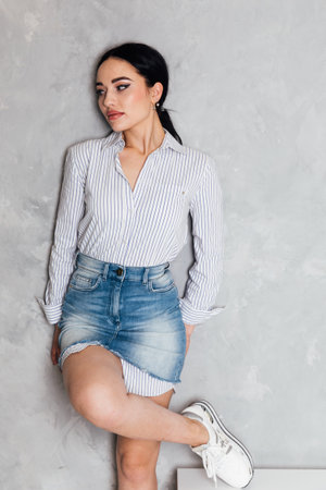Beautiful fashionable woman in denim skirt posing in studio on gray backgroundの写真素材