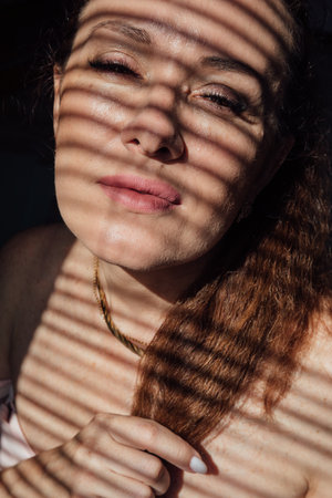 Beautiful woman 40 years old by the window with blinds light and shadowの写真素材