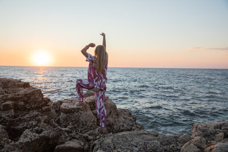 Blonde woman in silk suit on a rock by the sea at sunset hands upの写真素材