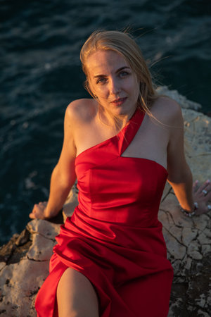 Blonde woman in red dress sitting on a rock by the seaの写真素材
