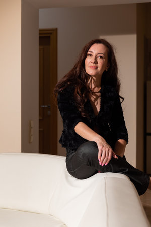Beautiful brunette woman in dark clothes sitting on white sofa in living room interiorの写真素材