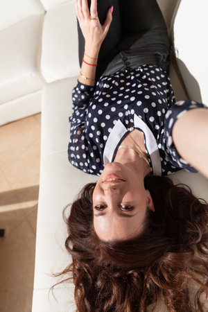 Beautiful woman in dark clothes lying on white sofa in living room interiorの写真素材
