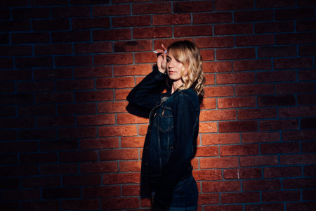Fashionable blonde woman in denim jacket and shorts standing against a brick wall