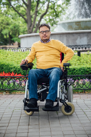 Smiling young disabled man use phone on a wheelchair in a park at sunlight outdoors.
