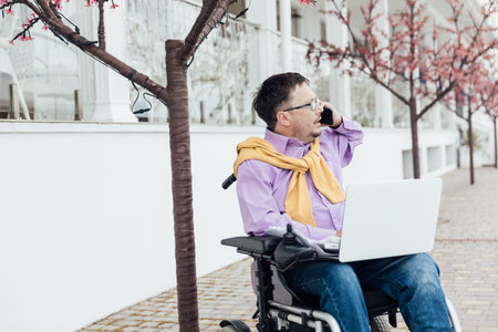 Young man in a wheelchair works at his laptop sitting against a beautiful sky.Disabled guy works at his laptop on the beach. Works remotely.の写真素材