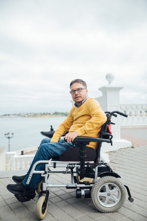 Smiling young disabled man use phone on a wheelchair in a park at sunlight outdoors. Disability cell...