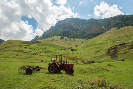 an old tractor without a driver with a rake stands in the valley, mountains, sun, paraglidersの写真素材