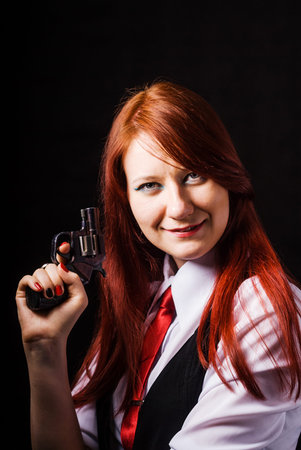 A red-haired beautiful girl in a white shirt, black vest and red tie, with a gun in her hand, looks at the camera, against a black background.の写真素材