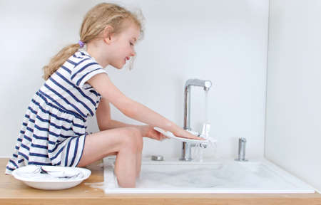 Happy little girl washing the dishes in the kitchen.の写真素材