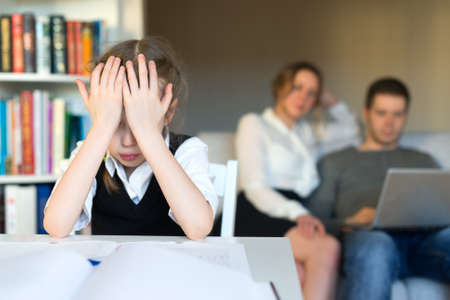 Upset little girl doing homework, while her parents resting on the sofa.の写真素材