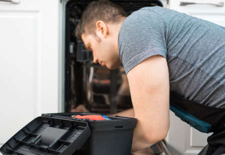 Professional handyman in overalls repairing domestic dishwasher in the kitchen.の写真素材