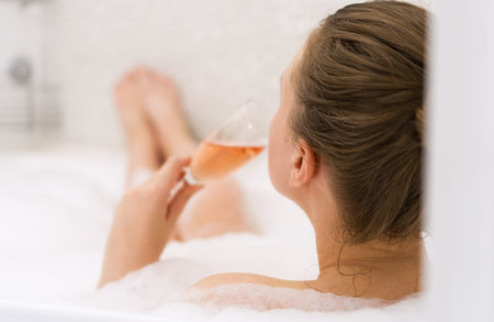Woman with glass of wine is relaxing in bathtub. Rear view.