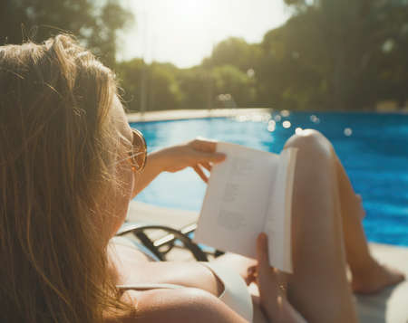 Pretty lady in sunglasses reading book near the pool in the summer.の写真素材