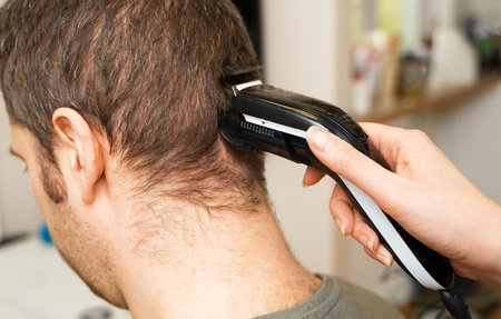Woman is doing haircut with hair trimmer to man at home.