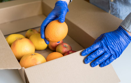 Volunteer in protective medical gloves putting fruits In donation box.