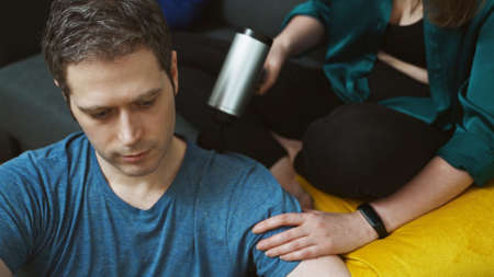 Woman massaging man with massage percussion device at home.の写真素材